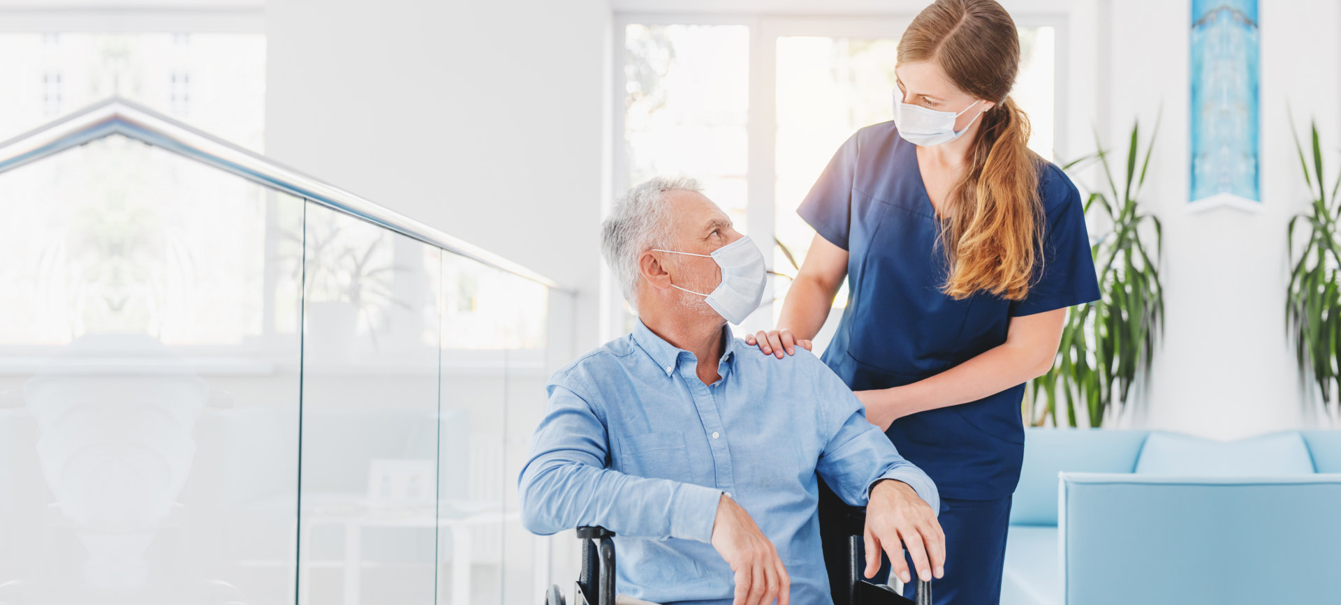 Young woman nurse explaining information to man patient in wheelchair in medical face mask while talking together in hospital.
